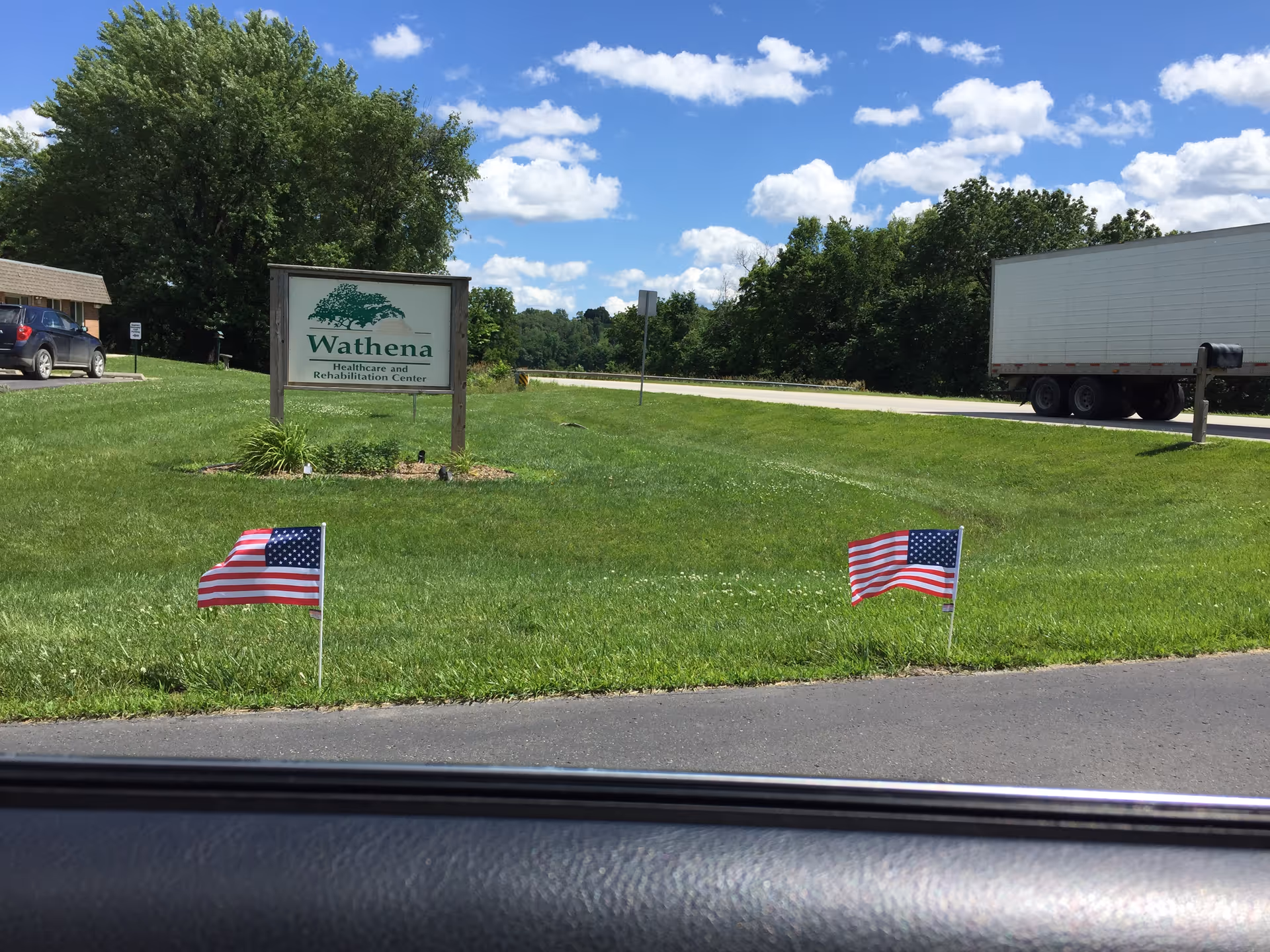 View of the grassy front lawn of Wathena Healthcare and Rehabilitation Center with a sign displaying the facility's name. Two small American flags are planted in the grass near the road. A large truck is parked on the road to the right, and a car is parked near a building on the left. Trees and a partly cloudy blue sky are visible in the background.