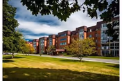 A multi-story brick senior living building with lawns and trees in the foreground under a partly cloudy sky.