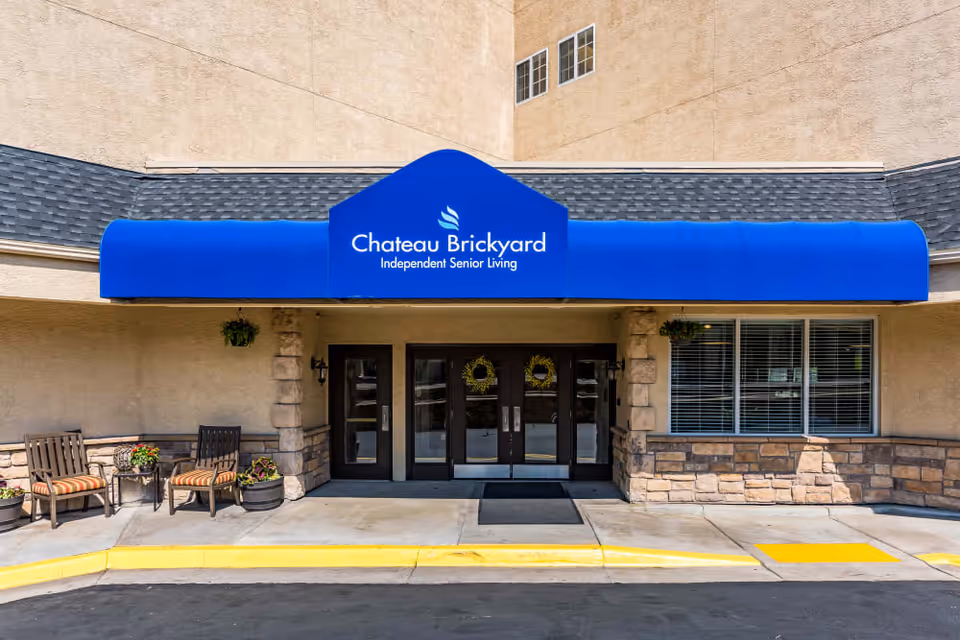Front entrance of a senior living building with a blue awning, double glass doors, and outdoor seating.