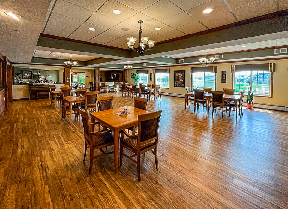 Spacious dining room with wooden floors and multiple wooden tables and chairs arranged neatly. Large windows on one side allow natural light to fill the room, and ceiling lights provide additional illumination. The room has a warm and inviting atmosphere with light-colored walls and decorative elements.