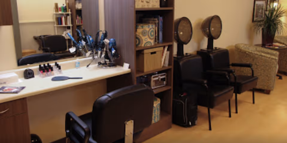 A salon area with styling chairs and hooded hair dryers next to a vanity mirror, countertop tools and shelving in a lounge-like room.