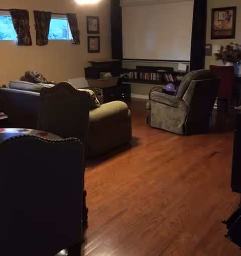 A cozy living room with wooden flooring featuring a sofa, an armchair, and a recliner. There are two small windows with curtains on the left wall, framed pictures on the walls, and a shelf with books and a projector screen on the far wall.