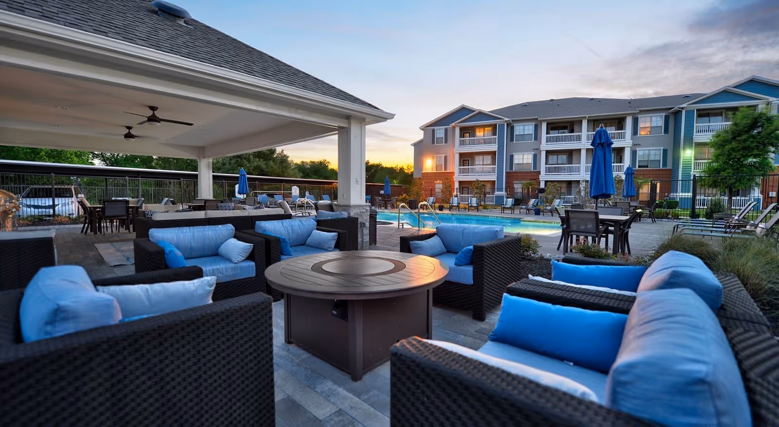 Outdoor seating area with black wicker chairs and blue cushions arranged around a round fire pit table, next to a covered patio with ceiling fans. In the background, there is a swimming pool, tables with umbrellas, and a multi-story residential building under a partly cloudy sky at sunset.