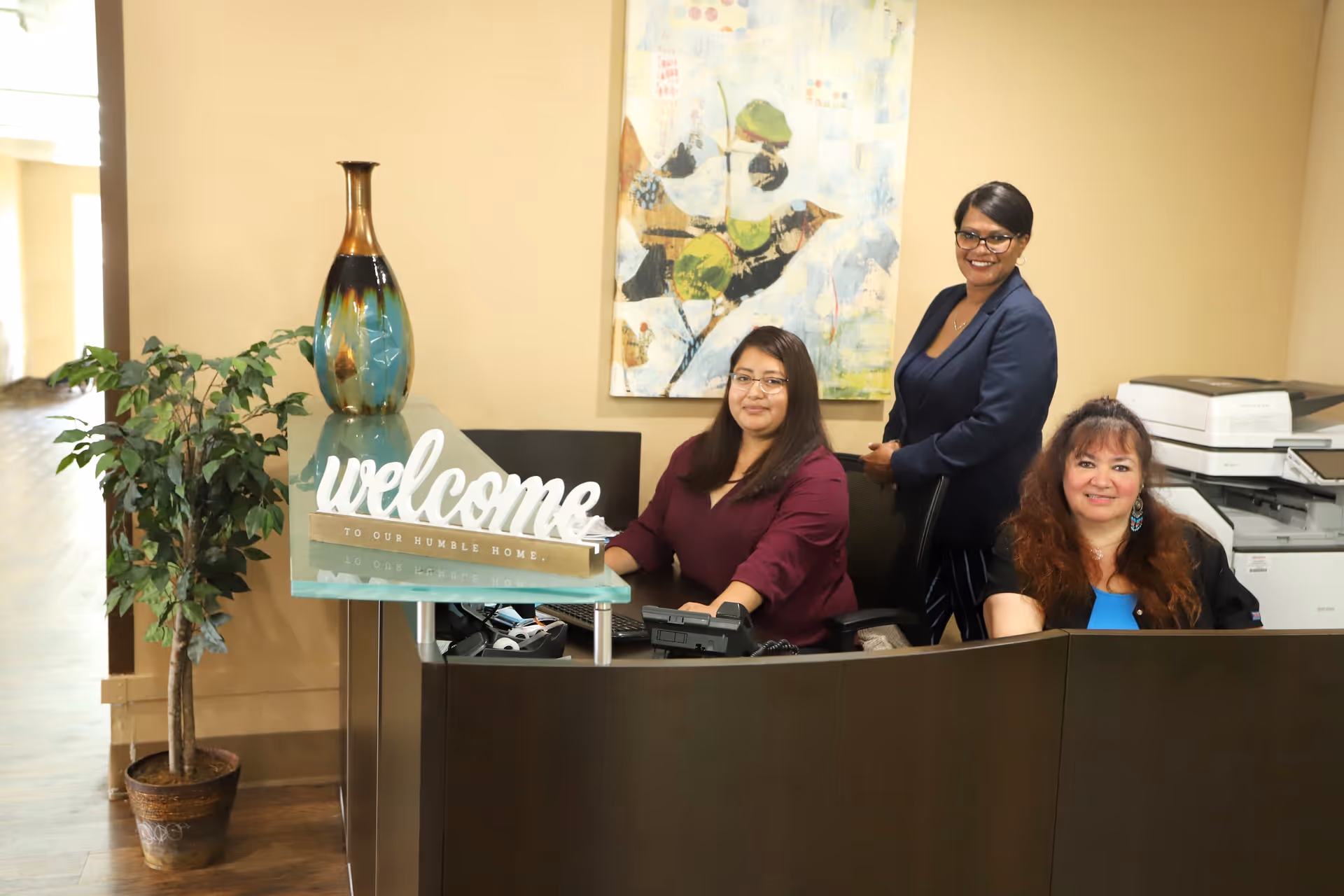 Three women at a reception desk in a facility. One woman is standing behind the desk while two women are seated. The desk has a decorative vase and a sign that says 'welcome to our humble home.' There is a potted plant to the left and a colorful painting on the wall behind them.