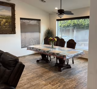 A dining room with a long covered wooden table and chairs, a vase of flowers, large windows, a ceiling fan, and wood-look flooring.