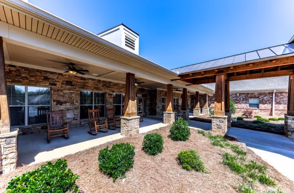 Outdoor covered patio area at Hope Center Memory Care with wooden rocking chairs, stone pillars, and landscaped bushes under a clear blue sky.