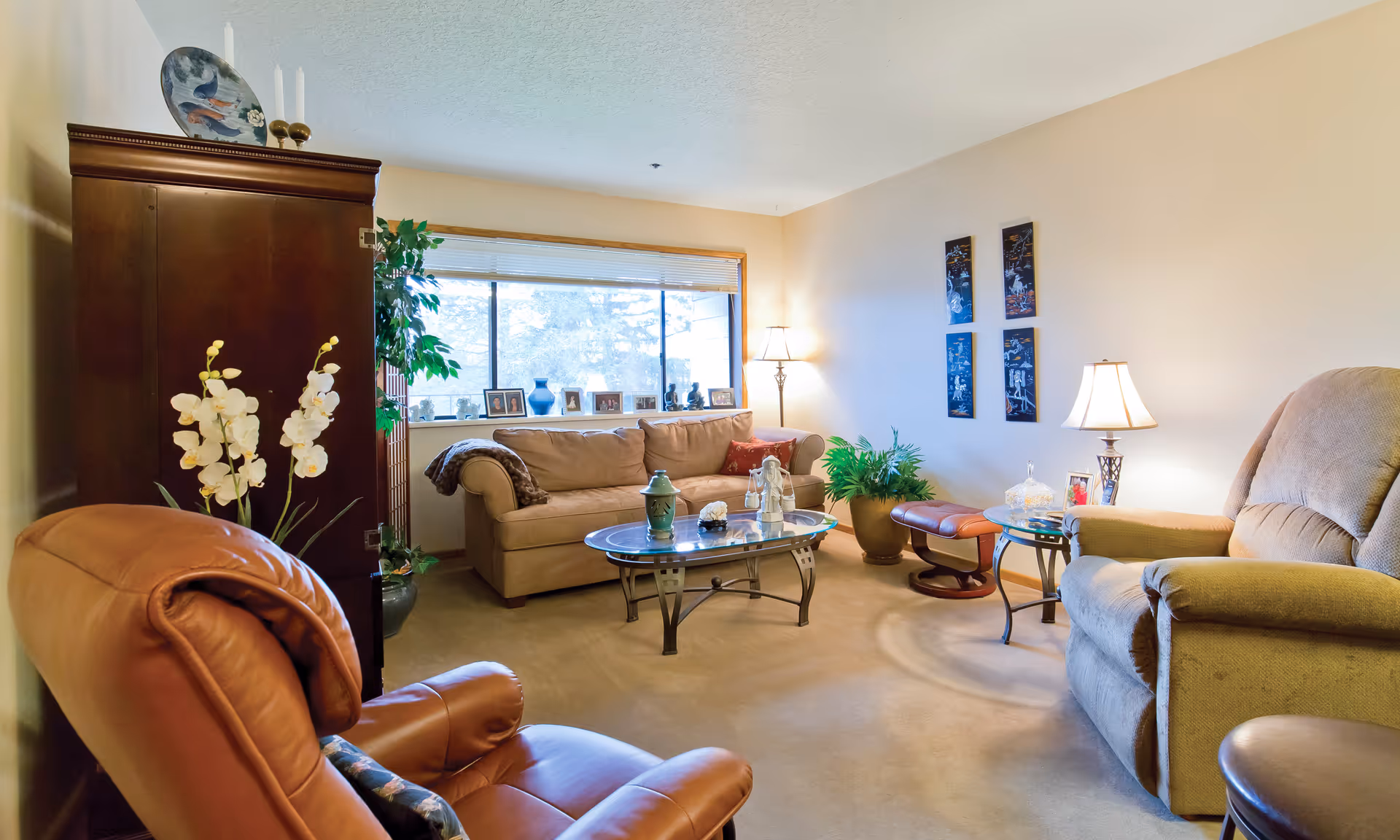 A cozy living room with a beige sofa, a brown leather recliner, and a beige armchair. There is a glass coffee table in the center with decorative items on it. The room has a large window with blinds, a wooden cabinet, potted plants, framed photos on the windowsill, and wall art. Two lamps provide warm lighting.