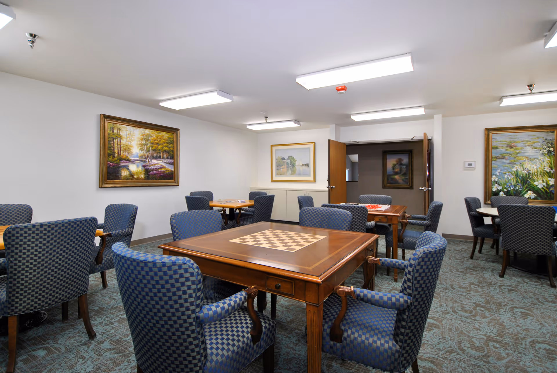 A well-lit room with multiple tables and blue checkered upholstered chairs arranged around them. The central table features a built-in chessboard. The walls are decorated with framed landscape paintings, and the carpet has a patterned design. The room appears to be a common area for activities or socializing.
