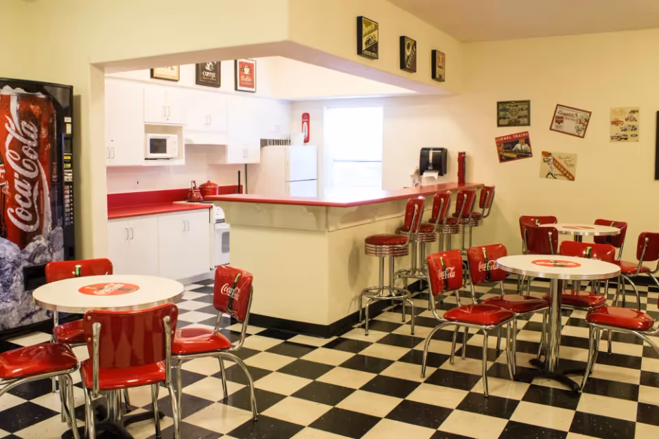 Retro Coca-Cola–themed dining area with red diner chairs and stools, round tables, a counter, checkered floor and a kitchenette in the back.