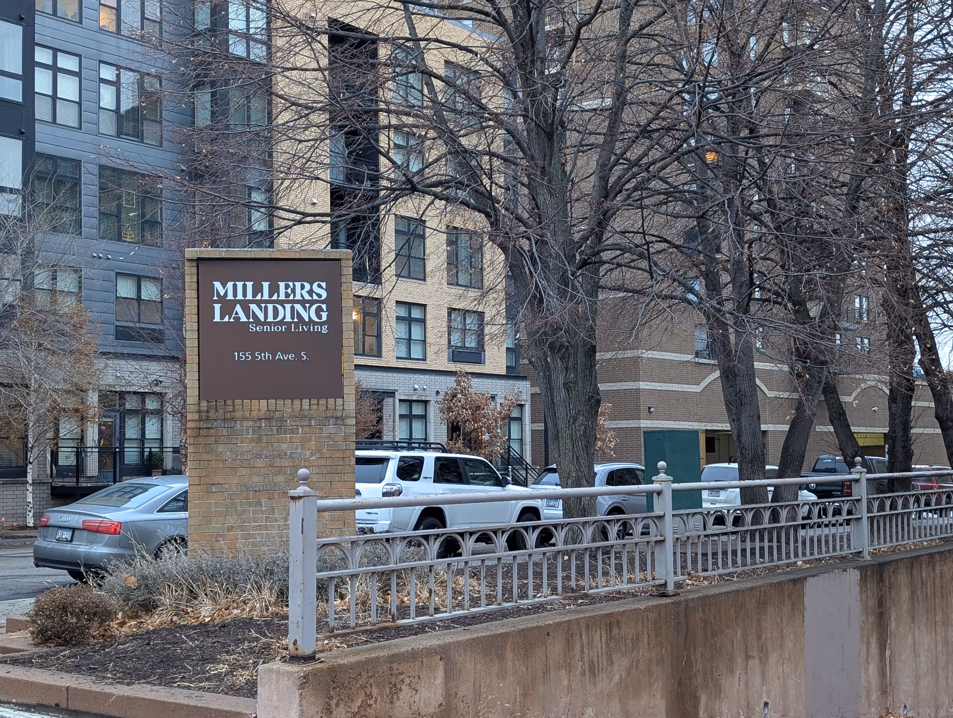 A brick sign reading "Millers Landing Senior Living" stands in front of a multi-story residential building with parked cars and leafless trees.