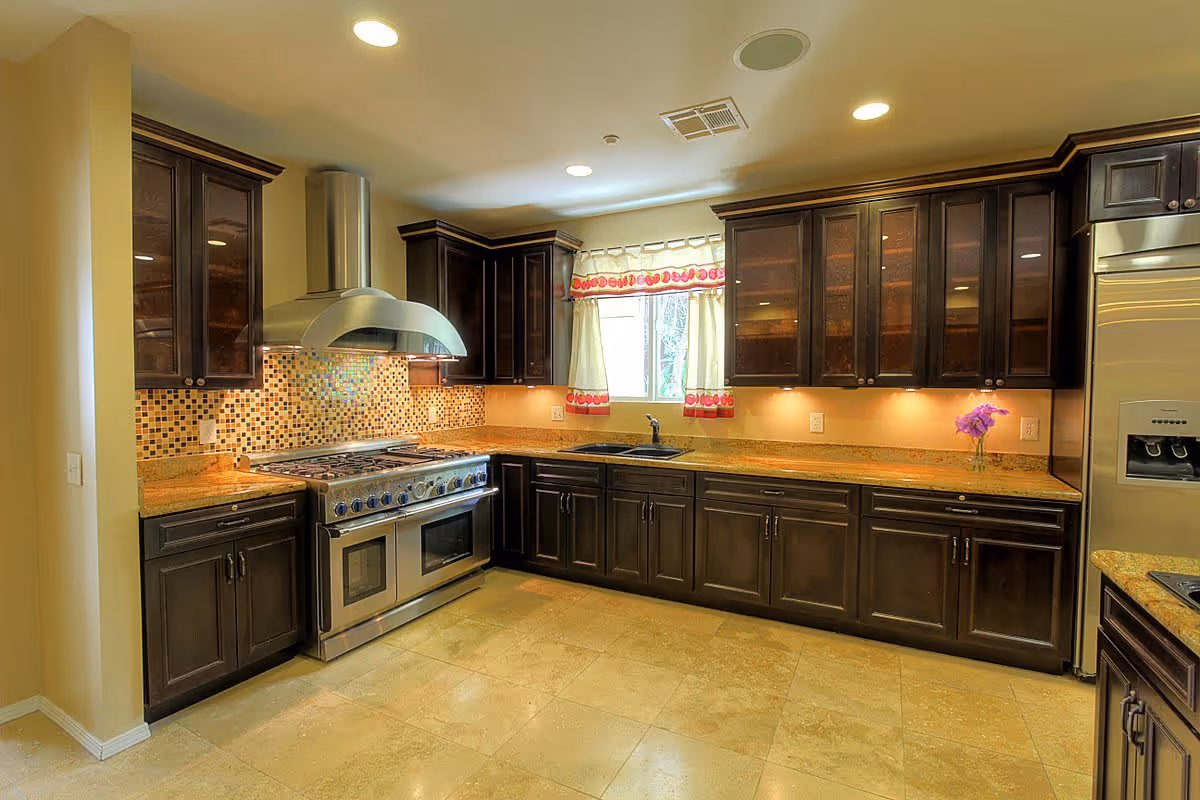 A modern kitchen with dark wooden cabinets, a stainless steel stove with a large range hood, a mosaic tile backsplash, granite countertops, a window with decorative curtains, and a stainless steel refrigerator with a water dispenser.