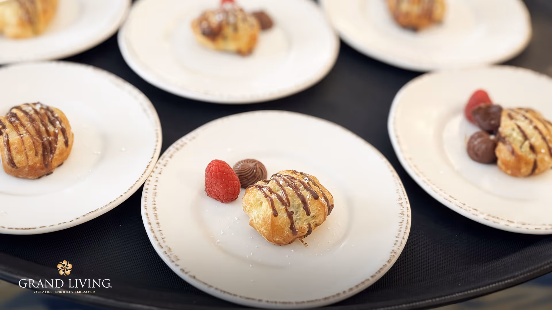 Several white plates each with a small pastry drizzled with chocolate, a raspberry, and a chocolate truffle, arranged on a black tray.