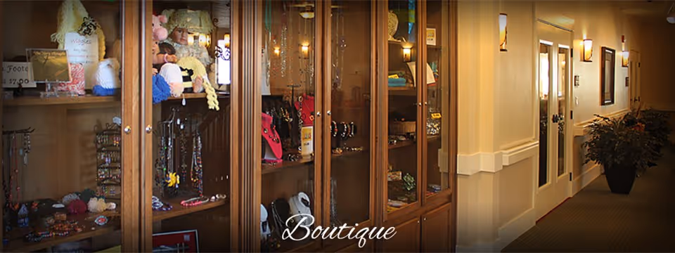 Interior view of a boutique area in a senior living facility featuring glass display cabinets filled with jewelry, accessories, and knitted items. The hallway adjacent to the boutique is softly lit with wall sconces and decorated with a large potted plant.