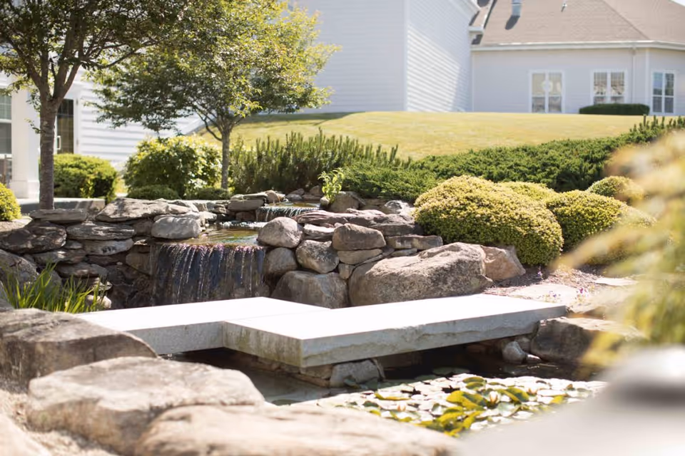 A serene outdoor garden area at StoneRidge Senior Living featuring a small stone waterfall, a stone bridge over a pond with lily pads, surrounded by bushes, trees, and a well-maintained lawn with a building in the background.