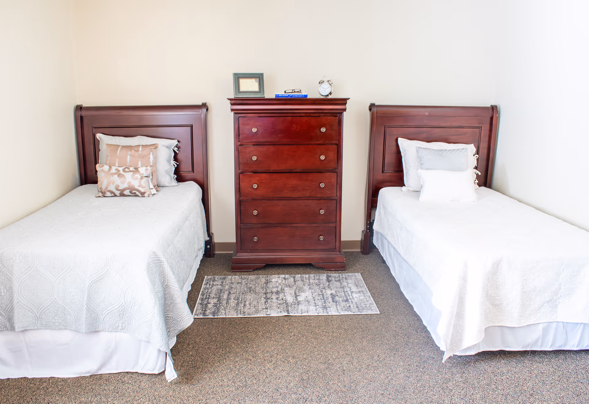 A simple bedroom with two twin beds separated by a dark wooden chest of drawers. Each bed has white bedding and decorative pillows. A small rug is placed on the carpeted floor in front of the chest of drawers. The walls are plain and light-colored.