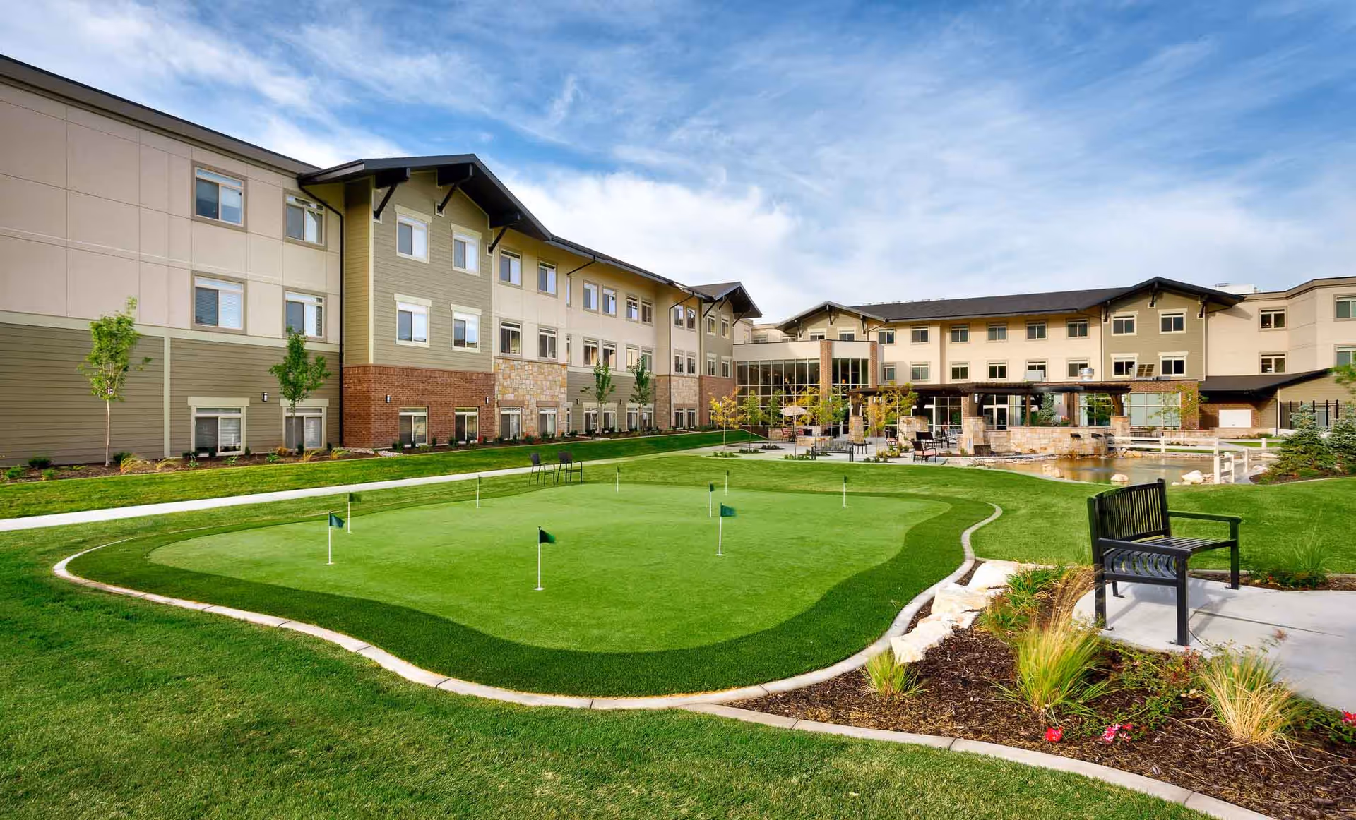 Outdoor view of Creekside Assisted & Senior Living facility showing a well-maintained putting green with several small flags, surrounded by landscaped grass and plants. The building has multiple windows and a mix of beige, green, and brick exterior finishes. There are benches and patio seating areas near a small pond under a partly cloudy sky.