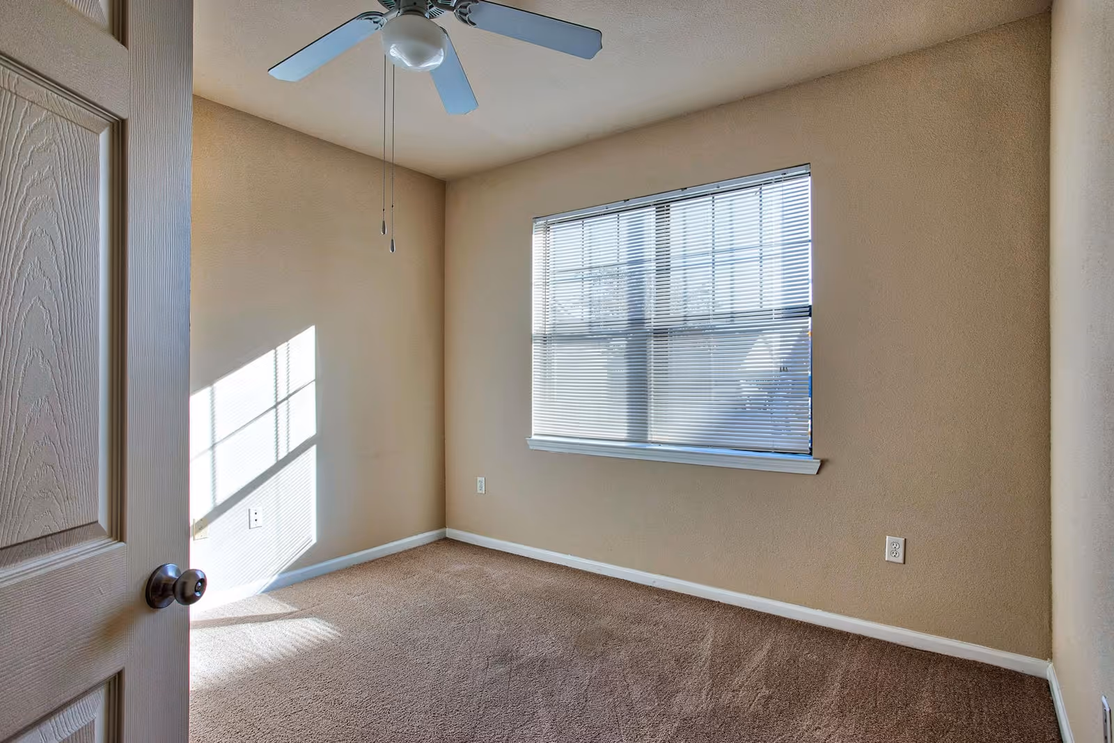 Empty room with beige walls and carpet, a window with closed blinds letting in sunlight, a ceiling fan with light, and a partially open door with a round knob.