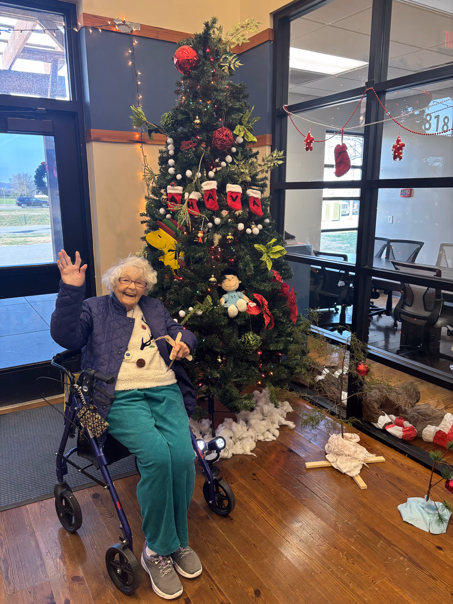 An elderly woman sitting on a walker and waving, smiling happily next to a decorated Christmas tree inside a facility. The tree is adorned with ornaments, small stockings spelling out 'NAVA', and festive decorations. The room has wooden floors, large windows, and office chairs visible through glass partitions.