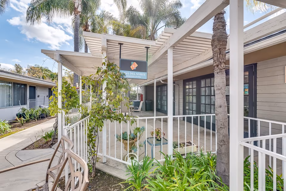 Outdoor covered walkway area at an assisted living facility with white railings and pergola-style roof. There are palm trees and green plants around the walkway. A sign hanging from the pergola reads 'Casa Del Norte Assisted Living.' The building has large windows and doors along the walkway.