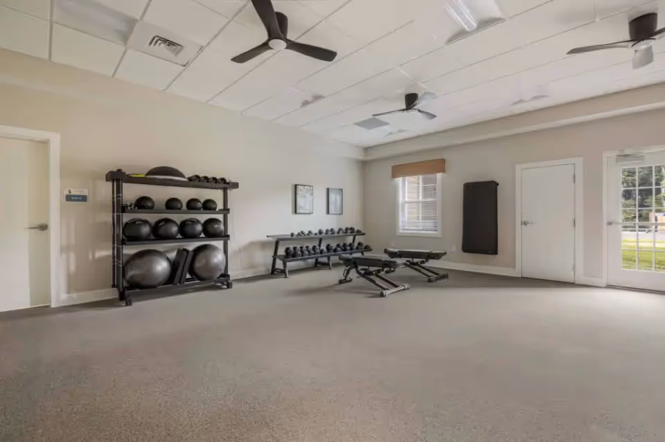 A fitness room with exercise equipment including medicine balls, dumbbells, two adjustable benches, and a wall-mounted exercise mat. The room has beige walls, a carpeted floor, ceiling fans, and windows letting in natural light.