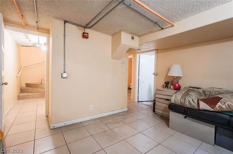 A small interior room with beige walls and tiled floor. There is a black couch with a blanket and pillows on the right side, next to a wooden nightstand with a lamp and some items. The room has exposed pipes on the ceiling and a doorway leading to a stairway and another room.