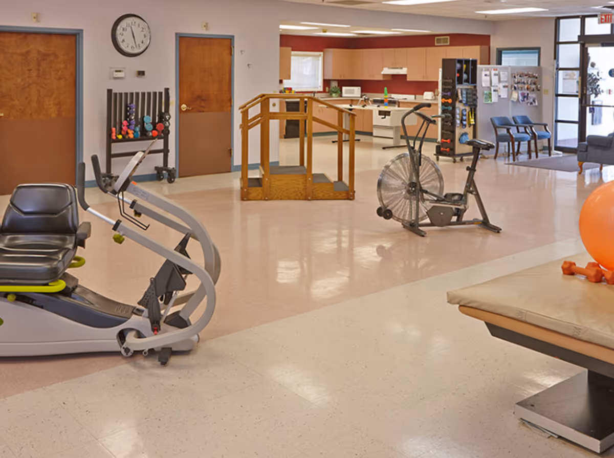 Interior view of a senior living facility exercise and rehabilitation area with exercise equipment including a recumbent bike, stationary bike, and a wooden staircase for physical therapy. There is a clock on the wall, a rack with colorful dumbbells, chairs near the entrance, and a kitchenette in the background.