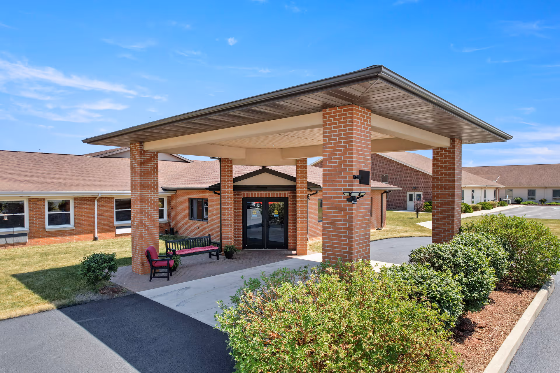 Entrance of a senior living facility with a covered drop-off area supported by brick columns. There are benches with red cushions placed near the entrance doors. The building is single-story with a brick exterior and a brown roof. Shrubs and a small lawn surround the driveway and entrance area under a clear blue sky.
