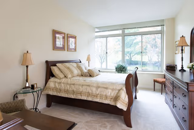 A bright bedroom with a large window letting in natural light. The room features a wooden bed with beige and cream striped bedding, two bedside tables with lamps, a wooden dresser with a mirror, a chair, and two framed pictures on the wall.