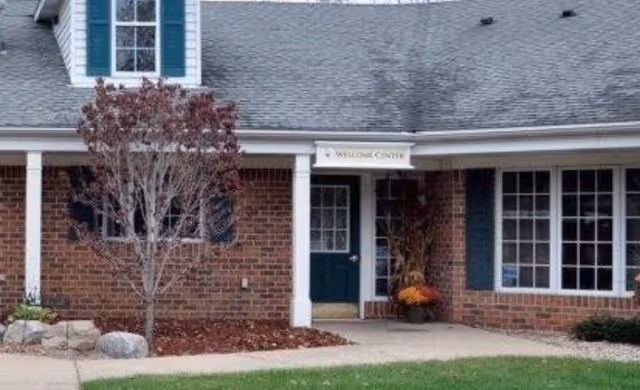 Exterior view of a brick building with a gray shingled roof and white trim. There is a small tree with red leaves in front of the building, a concrete walkway, and a sign above a door that reads 'Welcome Center'.