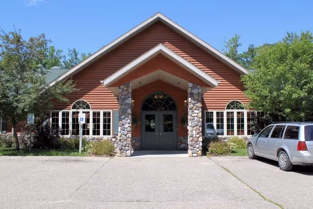 Front exterior view of a single-story building with a peaked roof, stone pillars framing the entrance, large windows on either side, surrounded by trees and bushes, with a silver car parked on the right side.