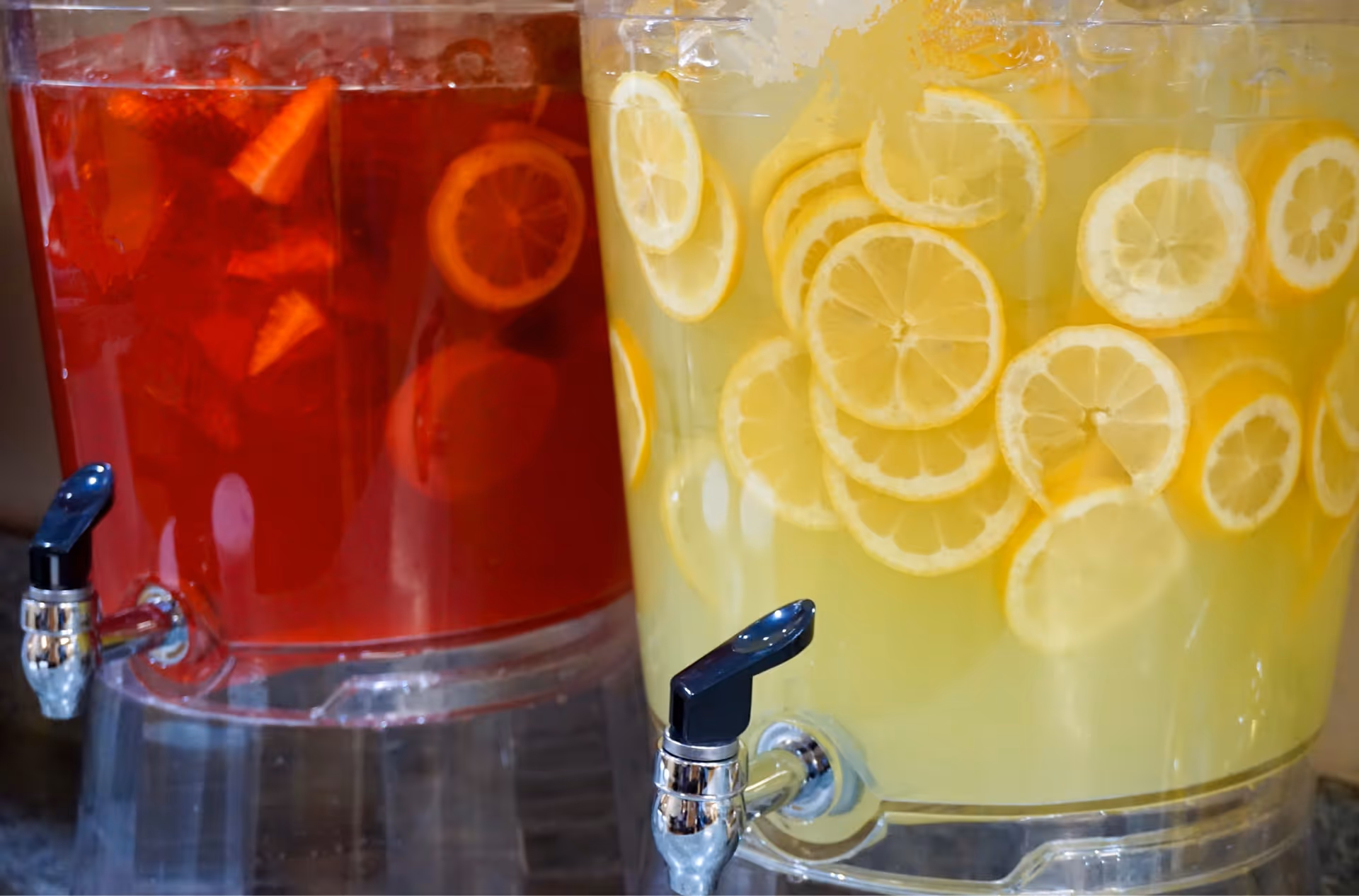 Two clear beverage dispensers on a counter, one filled with red fruit punch and the other with lemonade garnished with lemon slices.