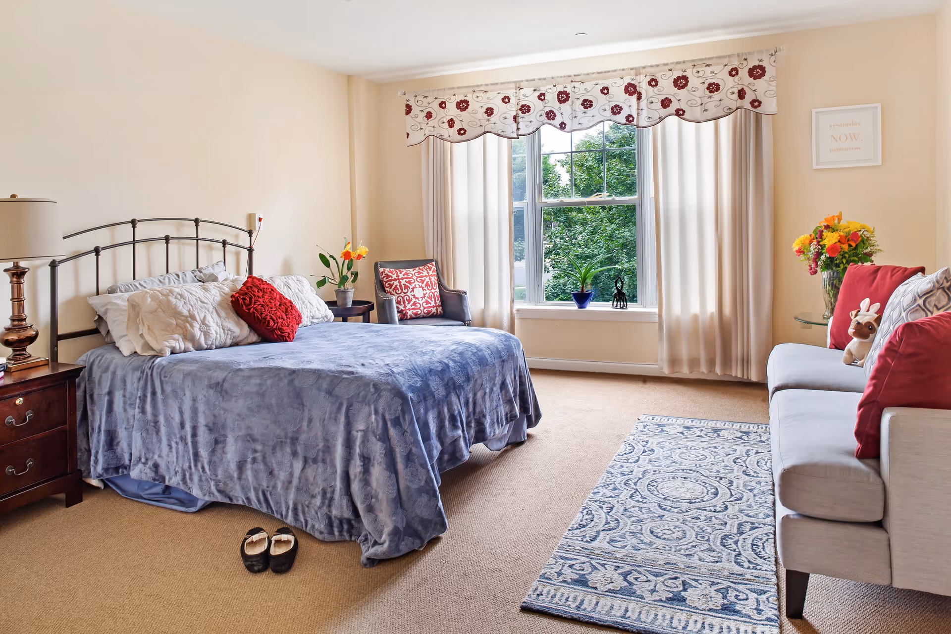 A cozy bedroom with a large bed covered in a blue blanket and multiple pillows, a wooden nightstand with a lamp, a patterned armchair with a red cushion, a window with floral valance and cream curtains, a small plant on the windowsill, a gray sofa with red and patterned cushions, a small stuffed animal, a glass side table with a vase of colorful flowers, and a blue patterned rug on the carpeted floor.