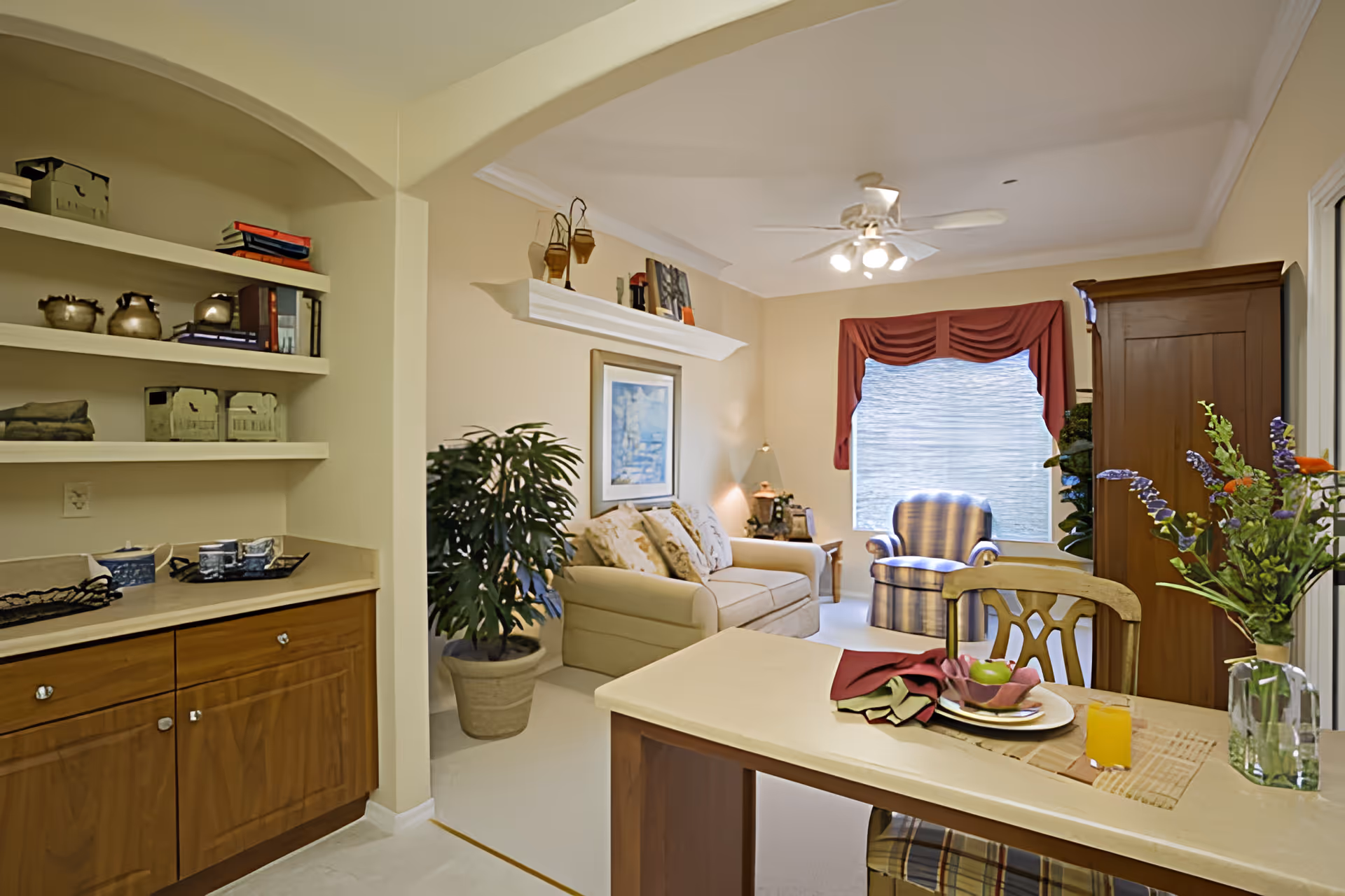 A cozy living room and dining area in a senior living facility. The room features a beige sofa with patterned cushions, a striped armchair near a window with red valance curtains, a wooden cabinet, and a ceiling fan with lights. A dining table with a chair is set with a plate, napkins, an apple, and a glass of orange juice. There are decorative plants and shelves with books and teapots on the left side.