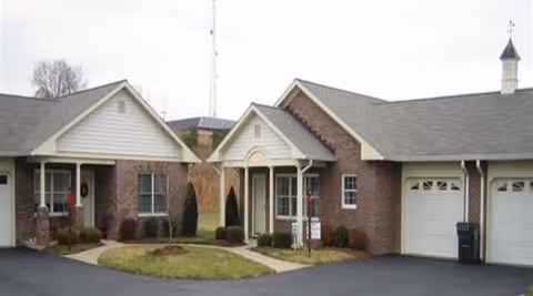 Exterior view of single-story brick and siding buildings with garages and small landscaped garden area in front, part of a senior living facility.