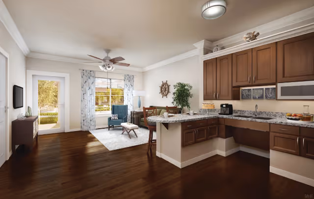 A bright and spacious senior living area featuring a kitchen with brown cabinets and granite countertops on the right, a small dining table with chairs in the center, and a cozy sitting area with two blue armchairs and a small ottoman near a large window with patterned curtains. A ceiling fan with lights is mounted on the ceiling, and a door leading outside is visible on the left. The room has dark hardwood floors and neutral-colored walls.