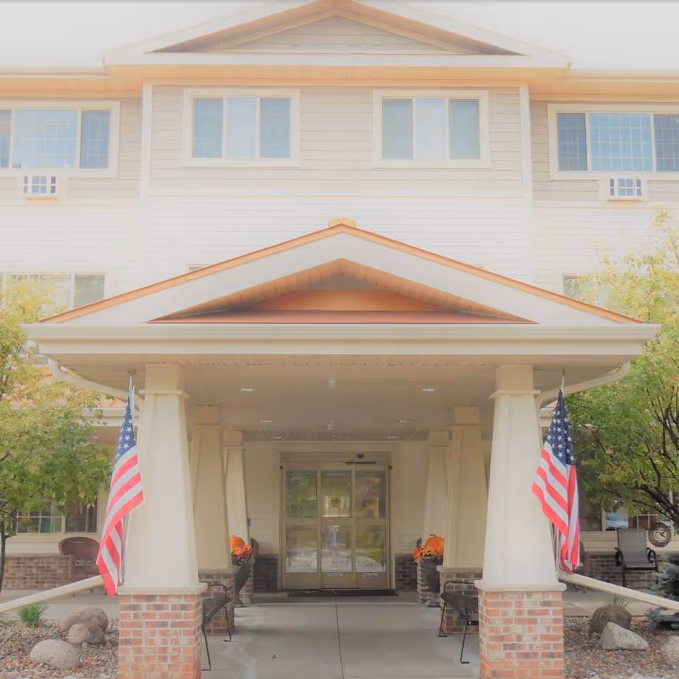 Front entrance of a multi-story assisted living facility with a covered porch supported by columns. Two American flags are displayed on either side of the entrance, with benches and potted plants nearby. Trees and landscaping surround the building.