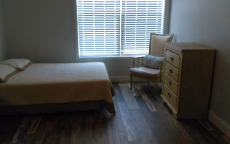 A simple bedroom with a bed covered in a beige blanket, a wooden dresser with four drawers, and a cushioned armchair with a pillow, all placed on a wooden floor. A window with white blinds is on the wall behind the chair and dresser.