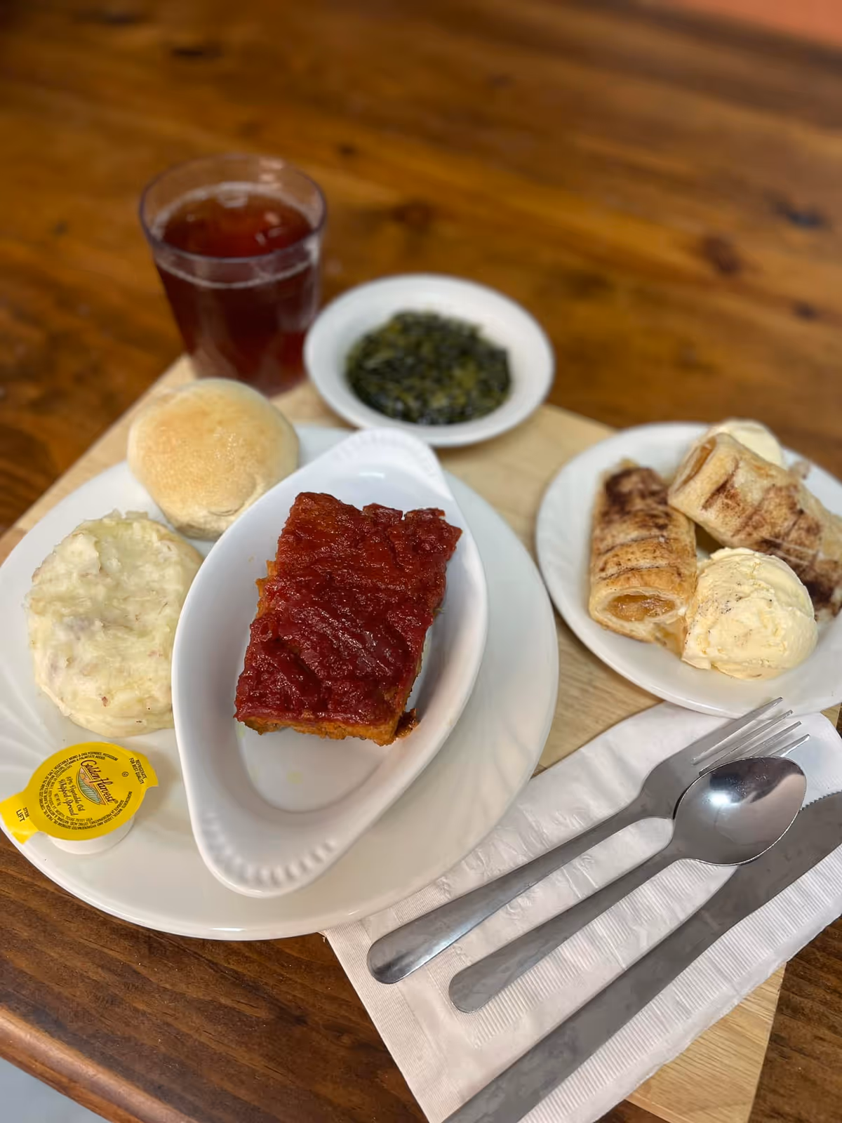 Plated meal on a wooden table featuring a slice of glazed meatloaf, mashed potatoes and a roll, a side of cooked greens, desserts and a glass of iced tea with utensils.