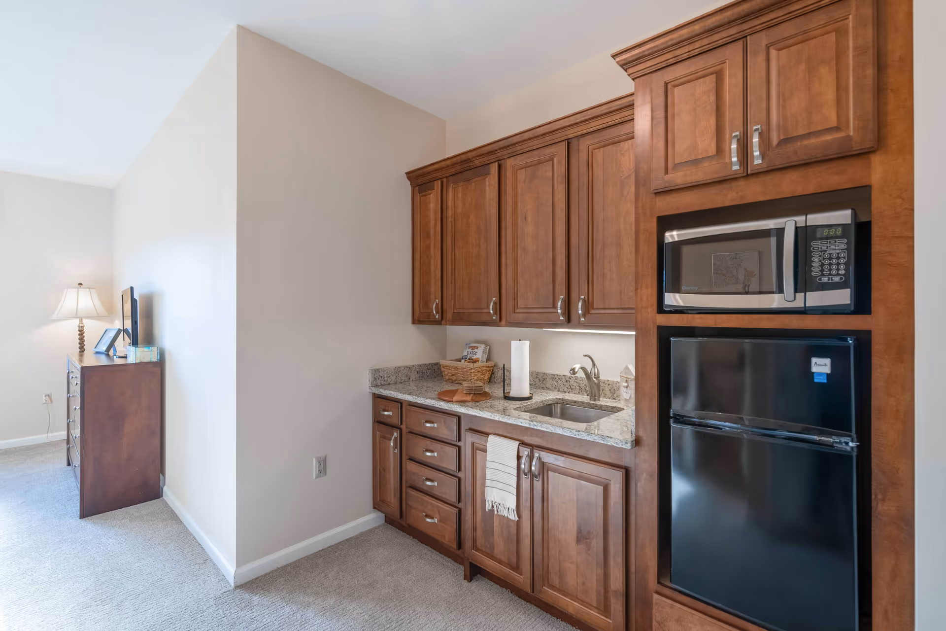 Small kitchenette with wooden cabinets, granite countertop, sink, microwave and mini-fridge in a senior living suite with a dresser and lamp visible in the adjacent room.