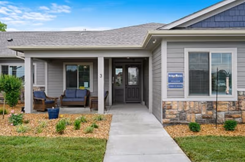 Front exterior view of a single-story residential building with a covered porch featuring outdoor seating. The building has gray siding with stone accents and a concrete walkway leading to a dark front door. There is a sign on the wall next to the window that reads Bridge Haven Village.