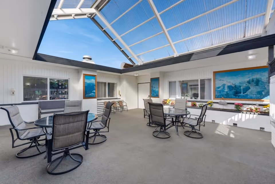 Indoor seating area with glass-top round tables and cushioned chairs under a large transparent skylight roof. The walls are white with framed landscape paintings and windows. There are flower arrangements on a ledge along one wall.