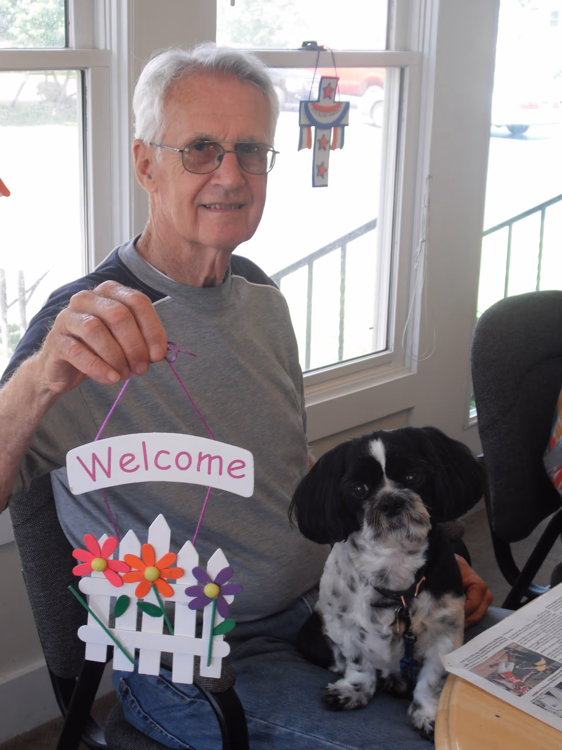 An elderly man with gray hair and glasses is sitting indoors holding a decorative sign that says 'Welcome' with colorful flowers on a white picket fence. A small black and white dog is sitting on his lap. There is a window behind them with a hanging patriotic decoration.