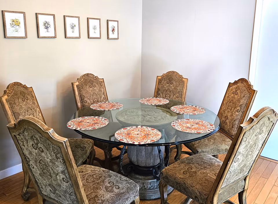 A round glass dining table with six ornate upholstered chairs arranged around it. The table has six round patterned placemats. On the wall behind the table, there are six framed floral artworks arranged in a horizontal line. The floor is wooden, and the room has light-colored walls.