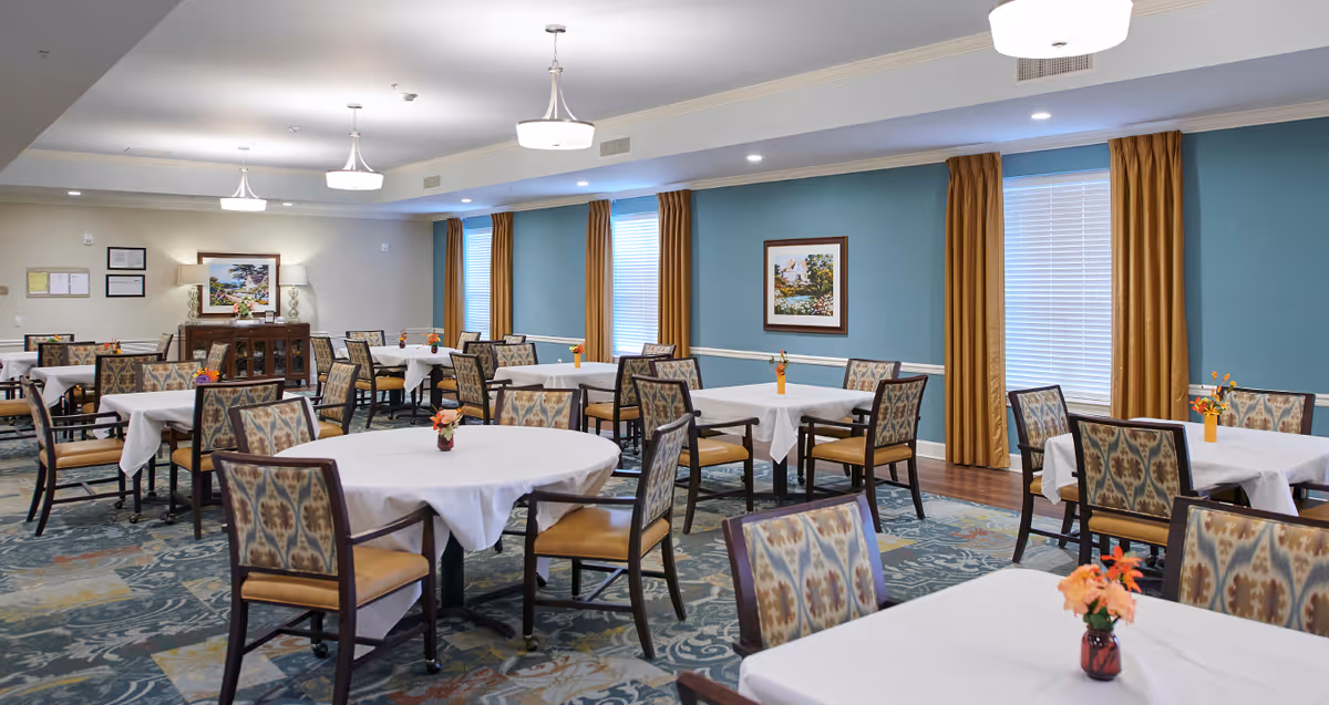 A spacious dining room with multiple tables covered in white tablecloths, each surrounded by patterned chairs. The room has blue walls with mustard-colored curtains on the windows, decorative paintings, and ceiling lights providing bright illumination. Small floral centerpieces are placed on each table.