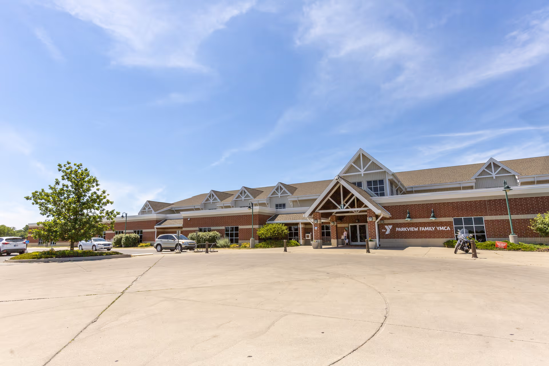 Exterior view of a large brick building with a peaked roof and multiple dormer windows under a blue sky. The building has a covered entrance with wooden beams and a sign that reads 'Parkview Family YMCA'. Several cars and a motorcycle are parked in front of the building on a concrete driveway.