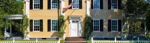 Two-story yellow building with white trim, black shutters, a flag, vines, and a white picket fence.