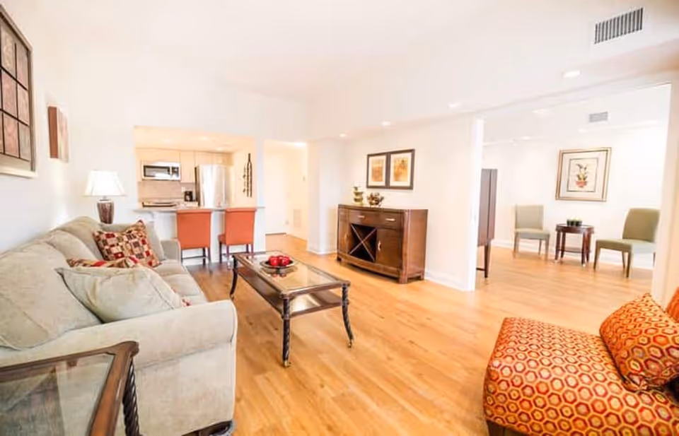 Bright and spacious living room with light wood flooring, a beige sofa with patterned cushions, a glass coffee table with red decorative items, an orange patterned armchair, and a wooden sideboard with framed artwork above it. The room opens into a kitchen area with a stainless steel refrigerator and two orange bar stools at the counter. In the background, there is a small seating area with two green chairs and a small table, decorated with framed botanical prints on the walls.