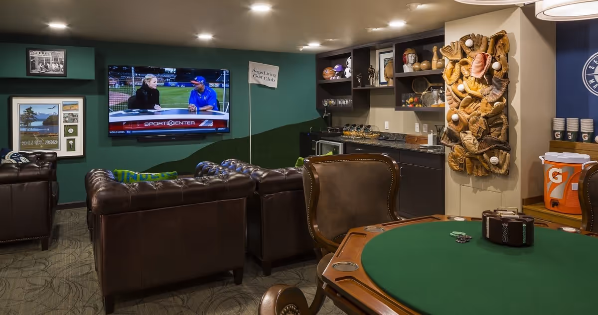 A cozy lounge area with dark leather sofas facing a wall-mounted TV showing a sports program. The room has green walls decorated with framed pictures and a small flag labeled 'Aegis Living Golf Club.' There is a poker table with green felt and leather chairs in the foreground. The back wall features a countertop with shelves holding various decorative items and a large wall art piece made of baseball gloves and balls. On the right side, there are Gatorade coolers and cups stacked on a counter.