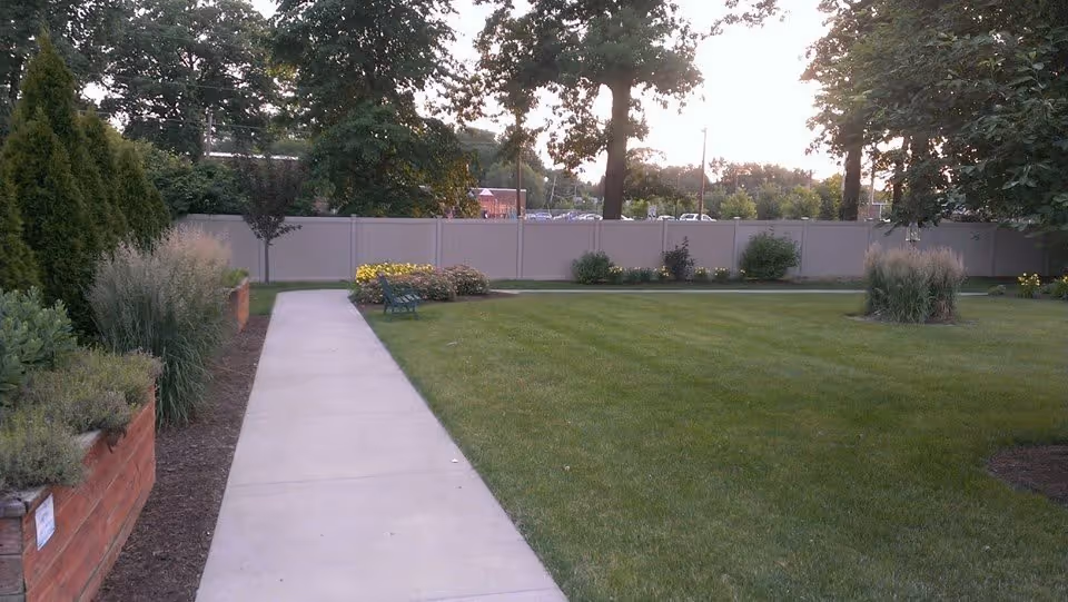 A well-maintained outdoor garden area with a concrete walkway, green grass, various shrubs, small trees, and a bench near the center. A tall fence and large trees are visible in the background under a bright sky.