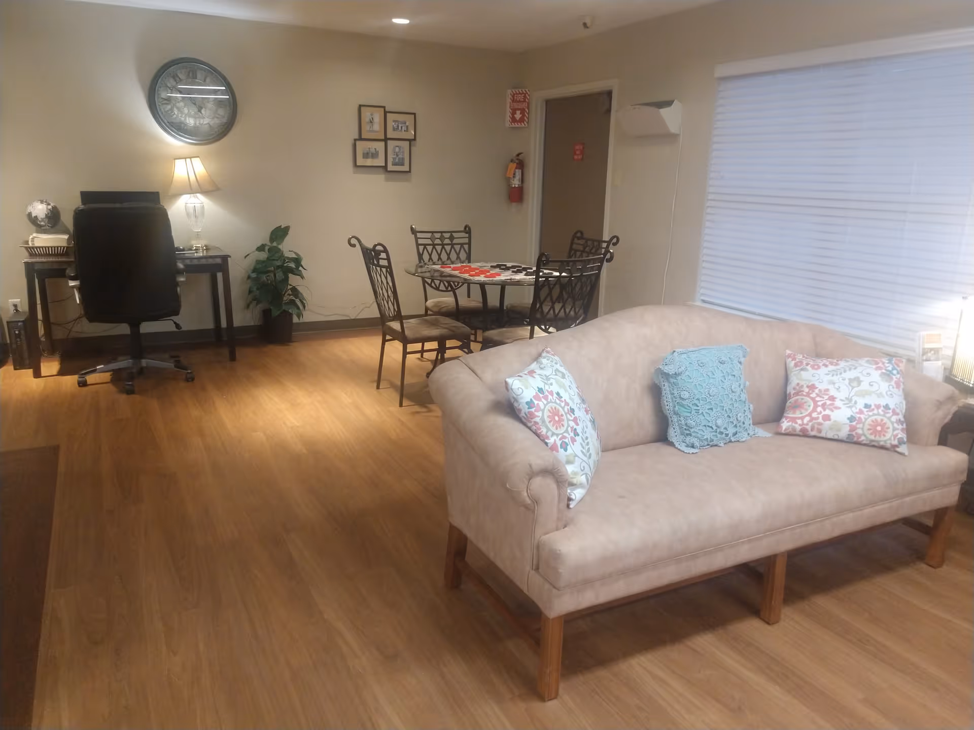 A cozy interior room with a beige couch adorned with three decorative pillows, a small round table with four chairs, a desk with a black office chair, a lamp, a wall clock, and framed pictures on the wall. The floor is wooden, and there is a window with closed blinds.